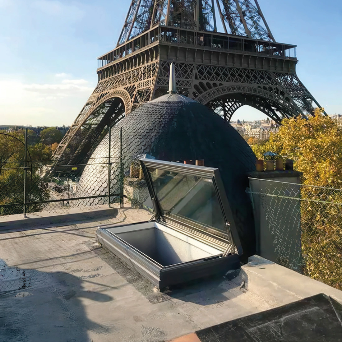 A view from a rooftop showing the Eiffel Tower in the background, with a dome-shaped structure and an open rooftop hatch in the foreground. Autumn trees are visible nearby under a clear blue sky.
