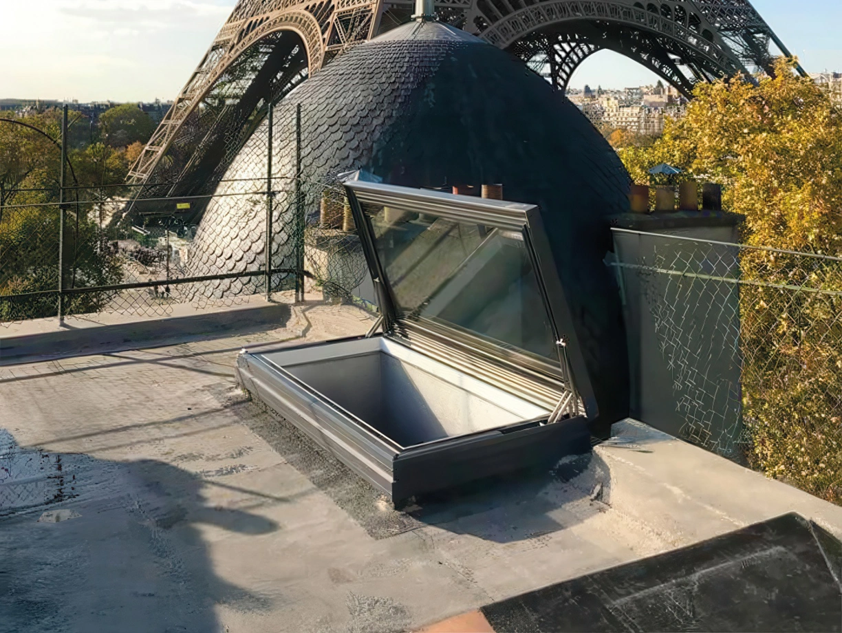Der Eiffelturm steht majestätisch im Hintergrund, während im Vordergrund ein geöffneter Dachluke sichtbar ist. Die Umgebung ist grün mit Bäumen und einem klaren blauen Himmel.