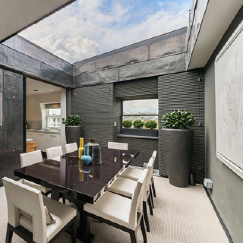 Comedor moderno con una mesa negra rectangular y sillas blancas. Las paredes son de metal oscuro y hay grandes ventanales. Un par de plantas decorativas y una obra de arte en la pared añaden un toque elegante.
