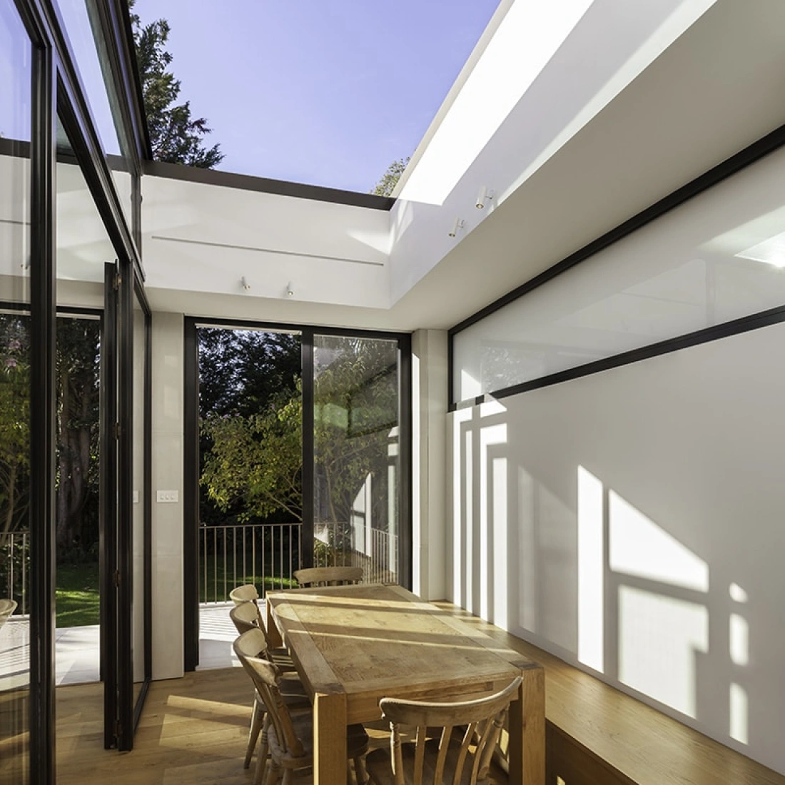 A modern dining area with a wooden table and four chairs, surrounded by large glass walls that let in natural light. The space features sleek design elements and looks out onto a green garden.