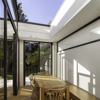 Salle à manger lumineuse avec une grande table en bois et quatre chaises, entourée de fenêtres ouvertes donnant sur un jardin. Des rayons de soleil illuminent le sol en bois.