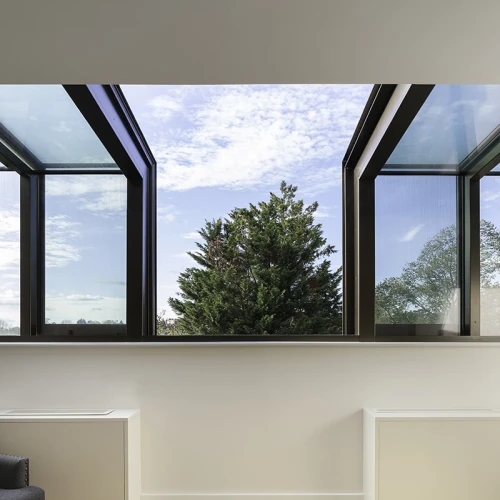 Ventana de un interior moderno con marcos de cristal abiertos, mostrando un árbol verde bajo un cielo azul con nubes. Suelo de madera clara.