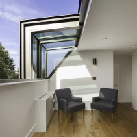 Two gray armchairs with wooden legs are positioned in a brightly lit area featuring a modern architectural design. Large windows fill the space with natural light, showing a blue sky outside. A sleek, minimalist wall and hardwood flooring complement the contemporary aesthetic.