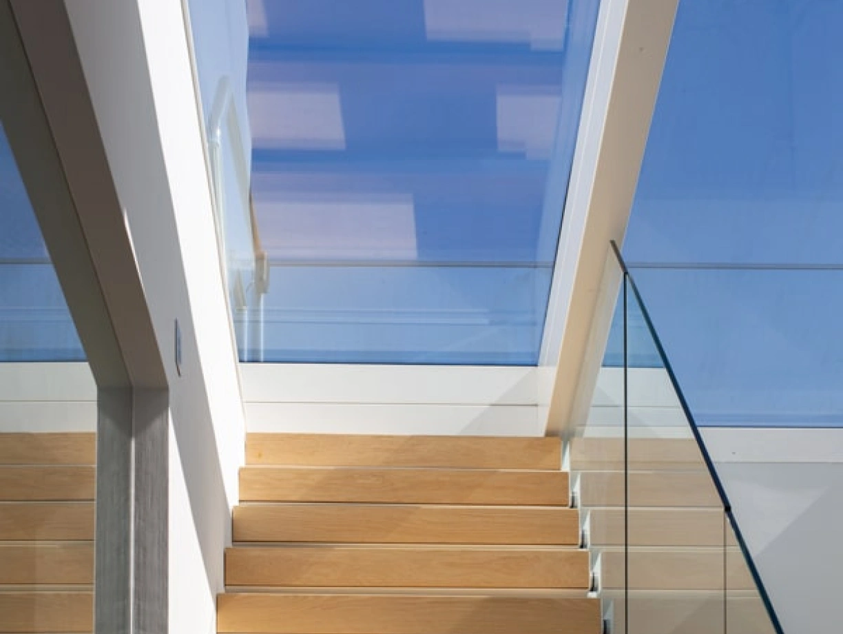 Escalera de madera clara con barandilla de vidrio, iluminada por una claraboya que deja entrar luz natural.