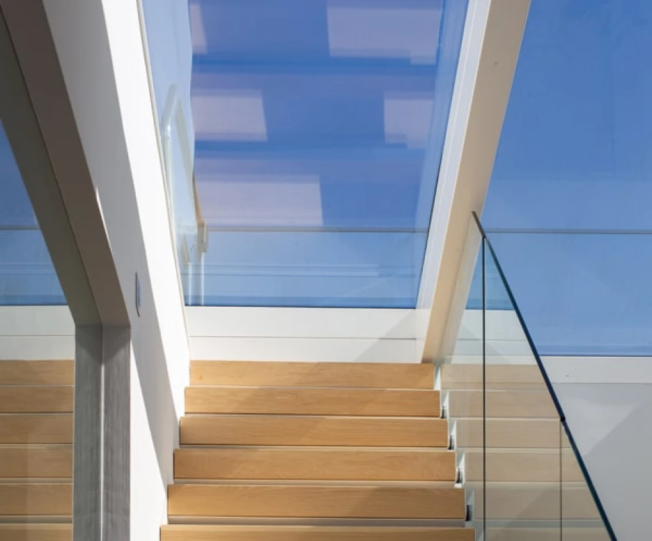 Escalera de madera clara con barandilla de vidrio, iluminada por una claraboya que deja entrar luz natural.