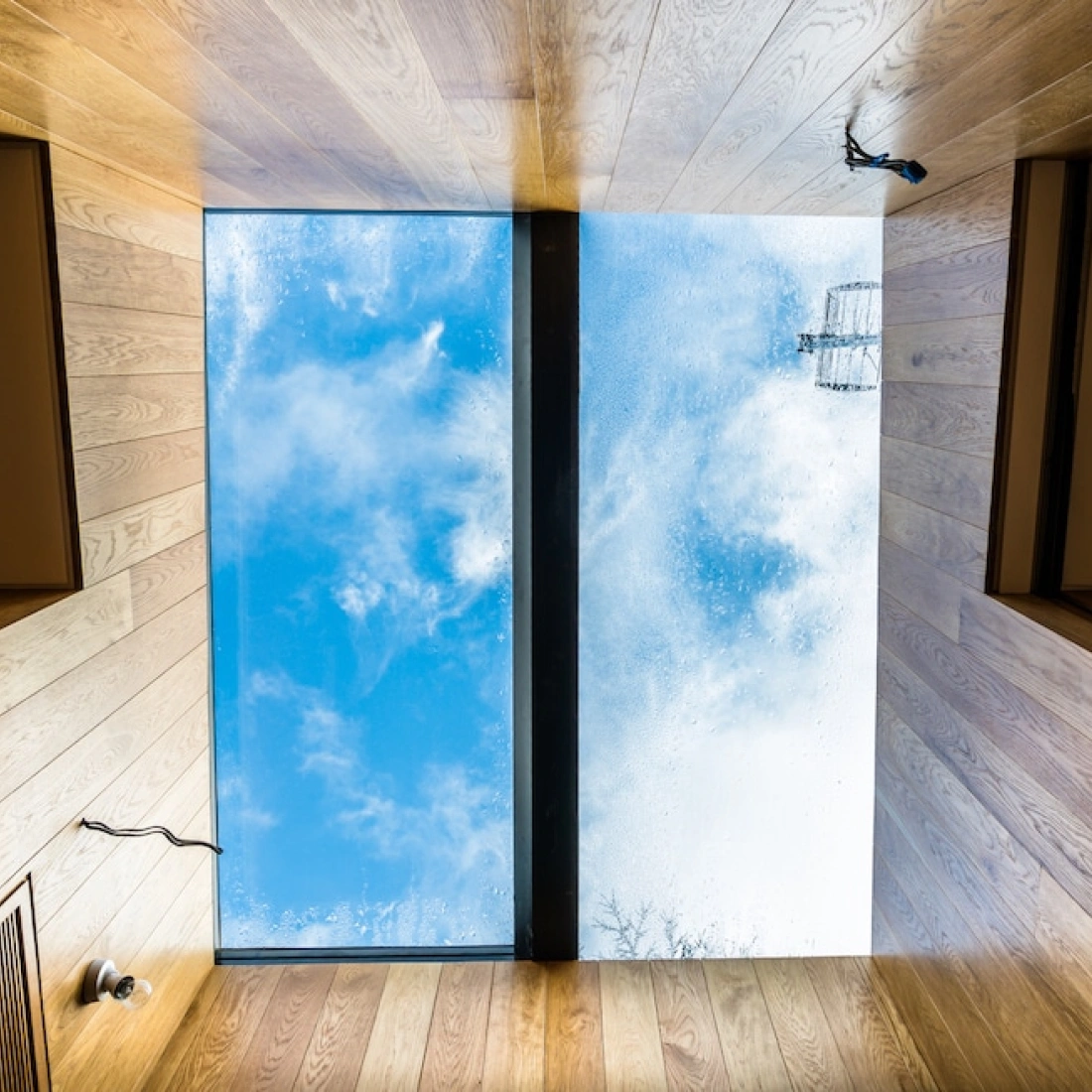 A flat ceiling with wooden panels and a large rectangular skylight showing a cloudy blue sky. Rainwater droplets are visible on the skylight's surface.