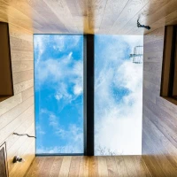 Vista desde el interior de una habitación con paredes de madera, mostrando una ventana rectangular en el techo que ofrece una vista del cielo nublado y algunos cables eléctricos.