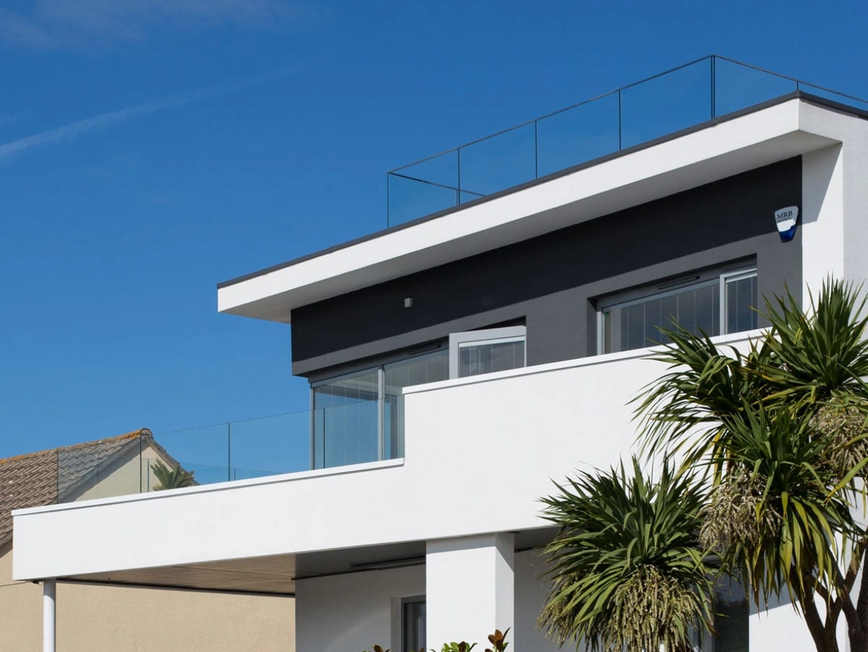 Modern two-story house with a flat roof and glass railing on the terrace. Clad in white and gray with large windows, surrounded by greenery under a clear blue sky.