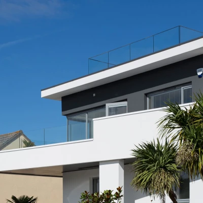 Modern two-story house with a flat roof and glass railing on the terrace. Clad in white and gray with large windows, surrounded by greenery under a clear blue sky.