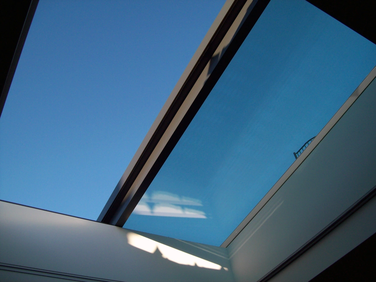 Visto desde el interior, un tragaluz muestra un cielo azul claro. La estructura del tragaluz es moderna, con bordes metálicos que contrastan con la suavidad del techo interior.