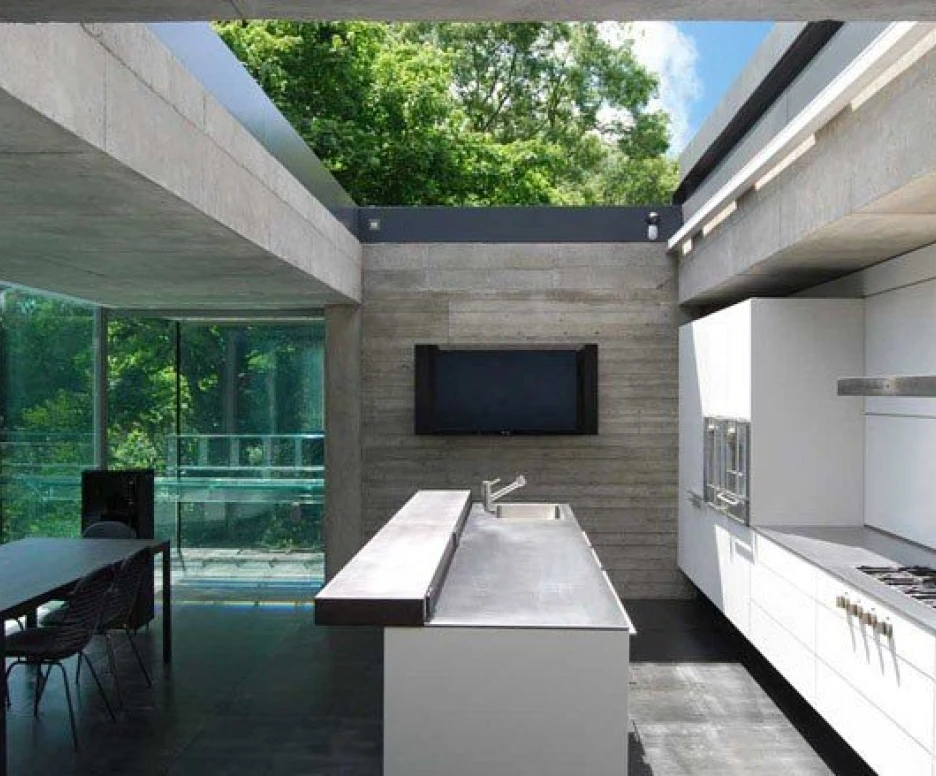 Cocina moderna con paredes de concreto, encimera de acero inoxidable y muebles blancos. Grandes ventanales ofrecen vistas al exterior arbolado. Una mesa y sillas se encuentran en el área del comedor.