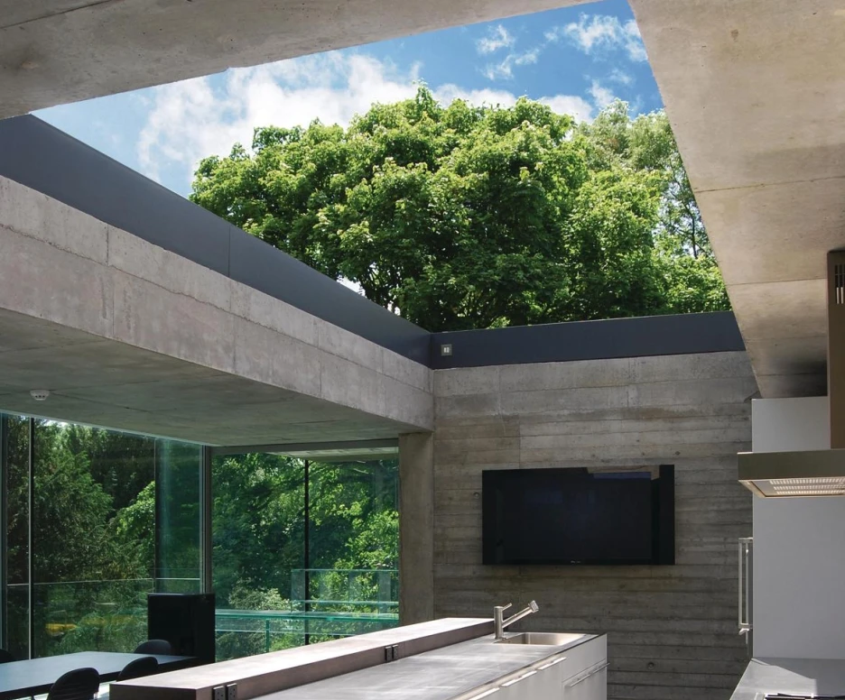 A modern kitchen featuring a sleek, white island with drawers, large windows allowing natural light, and a view of green trees outside. Neutral-colored walls and flooring complement the minimalist design, with a wall-mounted television in the background.