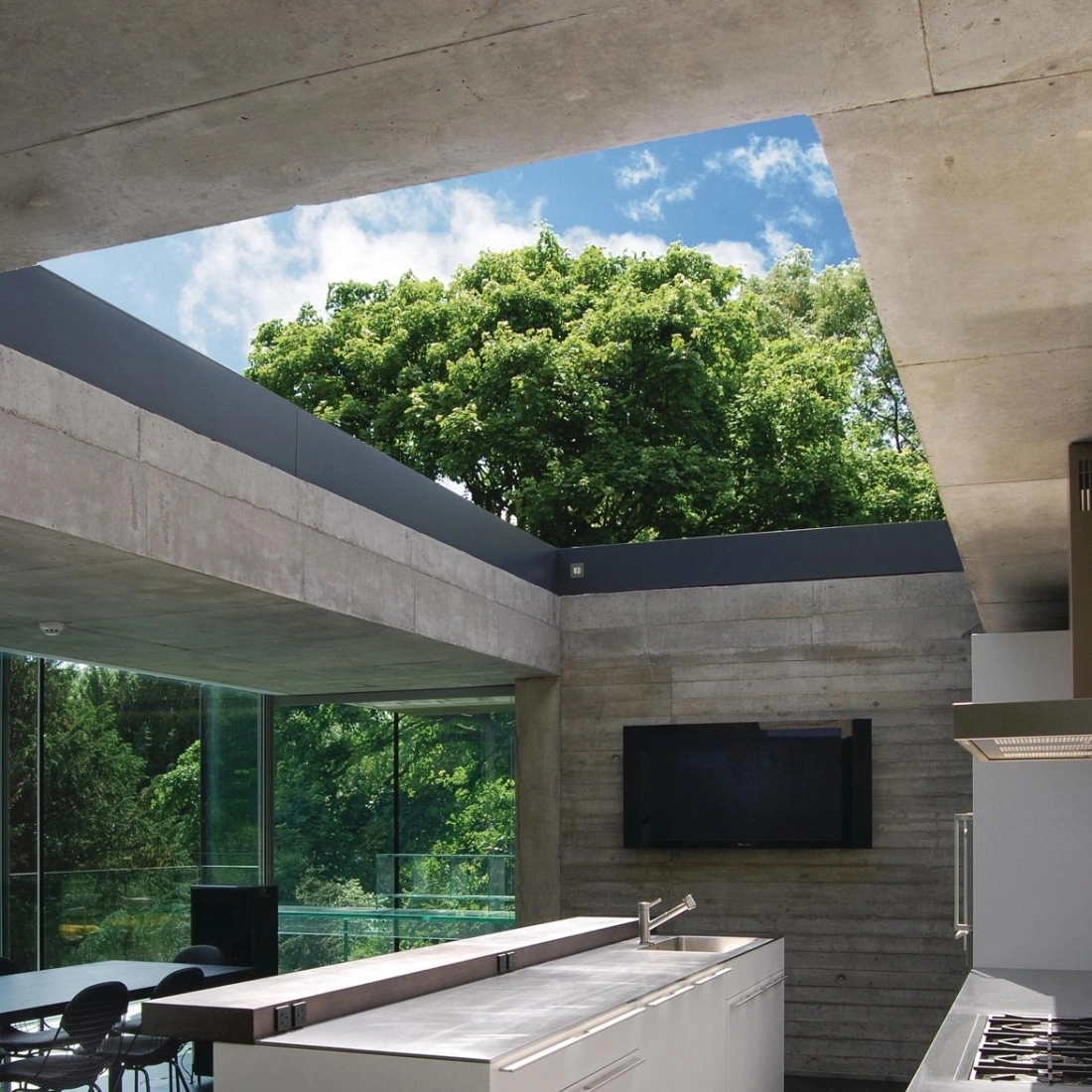 Modern kitchen featuring a contrasting white island with sleek cabinetry, large windows, and a view of lush greenery. The ceiling showcases concrete elements, and a flat-screen TV is mounted on the wall.