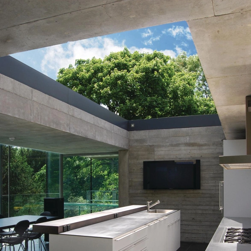 A modern kitchen featuring a sleek, white island with drawers, large windows allowing natural light, and a view of green trees outside. Neutral-colored walls and flooring complement the minimalist design, with a wall-mounted television in the background.