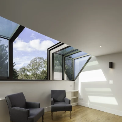 Espacio interior iluminado con dos sillones grises frente a una ventana grande con vidrio azul. El suelo es de madera clara y hay estanterías en la pared. Se pueden ver árboles y un cielo parcialmente nublado a través de la ventana.