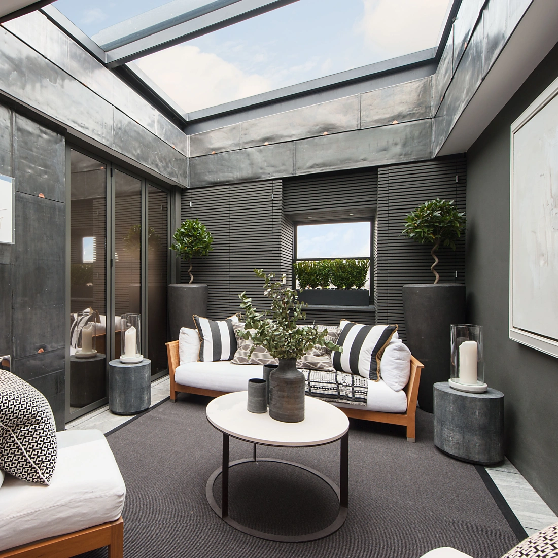 Espacio interior moderno con paredes de metal oscuro, sofá blanco y cojines decorativos. Hay una mesa de café circular y plantas en macetas. Una gran claraboya proporciona luz natural. Este ambiente combina elegancia y confort.