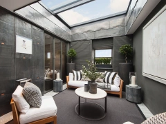 Espacio interior moderno con paredes de metal oscuro, sofá blanco y cojines decorativos. Hay una mesa de café circular y plantas en macetas. Una gran claraboya proporciona luz natural. Este ambiente combina elegancia y confort.