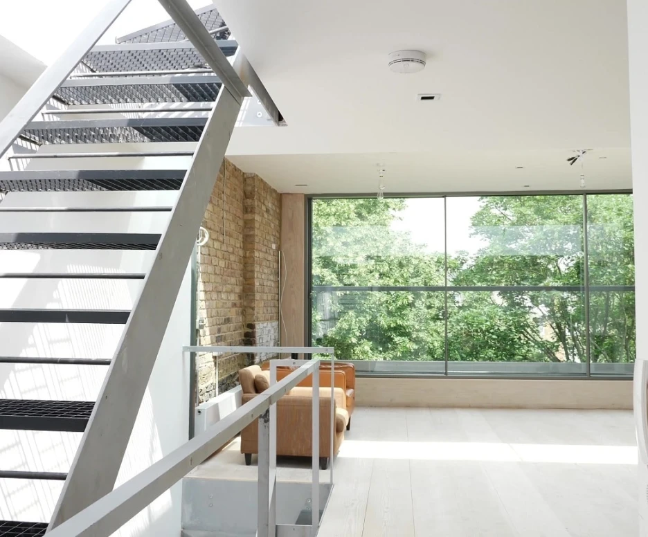 A modern interior featuring a metal staircase leading to an upper level. Large windows showcase trees outside, and a cozy seating area is visible. Natural light floods the space, highlighting the minimalist design.