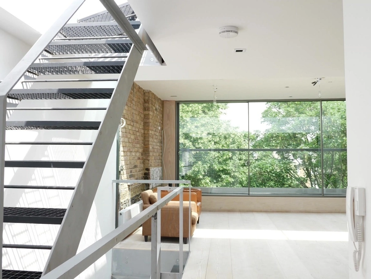 A modern staircase with a metal framework leads up to a bright, airy living space featuring large windows that overlook greenery. The interior has a combination of exposed brick and sleek white walls, with a cozy seating area visible in the background.