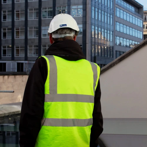 Ein Mann mit grauem Haar trägt eine gelbe Sicherheitsweste und einen weißen Helm. Er steht auf einem Balkon und blickt auf moderne Bürogebäude.