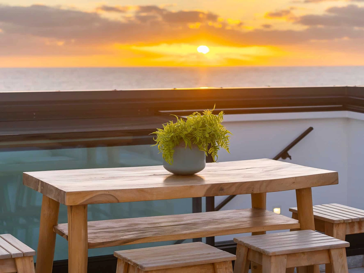A wooden table with a potted fern sits on a rooftop terrace, overlooking a sunset over the ocean. The sky is painted with warm hues of orange and yellow.