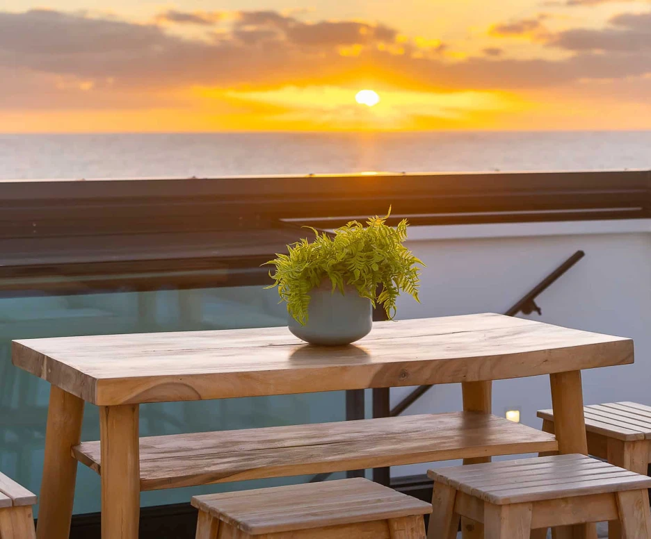 Un coucher de soleil sur l’océan, vu depuis un balcon avec une table en bois et des bancs. Un pot de plante verte est placé au centre de la table.