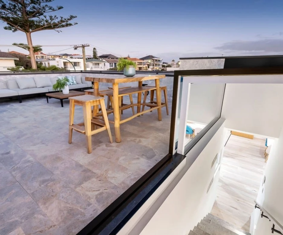 A modern rooftop terrace featuring a large wooden dining table with stools, surrounded by comfortable seating. The area is adorned with potted plants and overlooks a neighborhood with houses in the distance, under a twilight sky. A staircase leads down from the terrace.