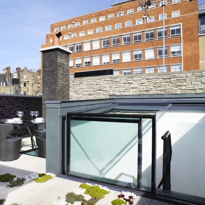 A modern rooftop terrace features a glass canopy, a table set for two with black chairs, and greenery among stone and wooden elements. In the background, a multi-story brick building is visible against a clear blue sky.