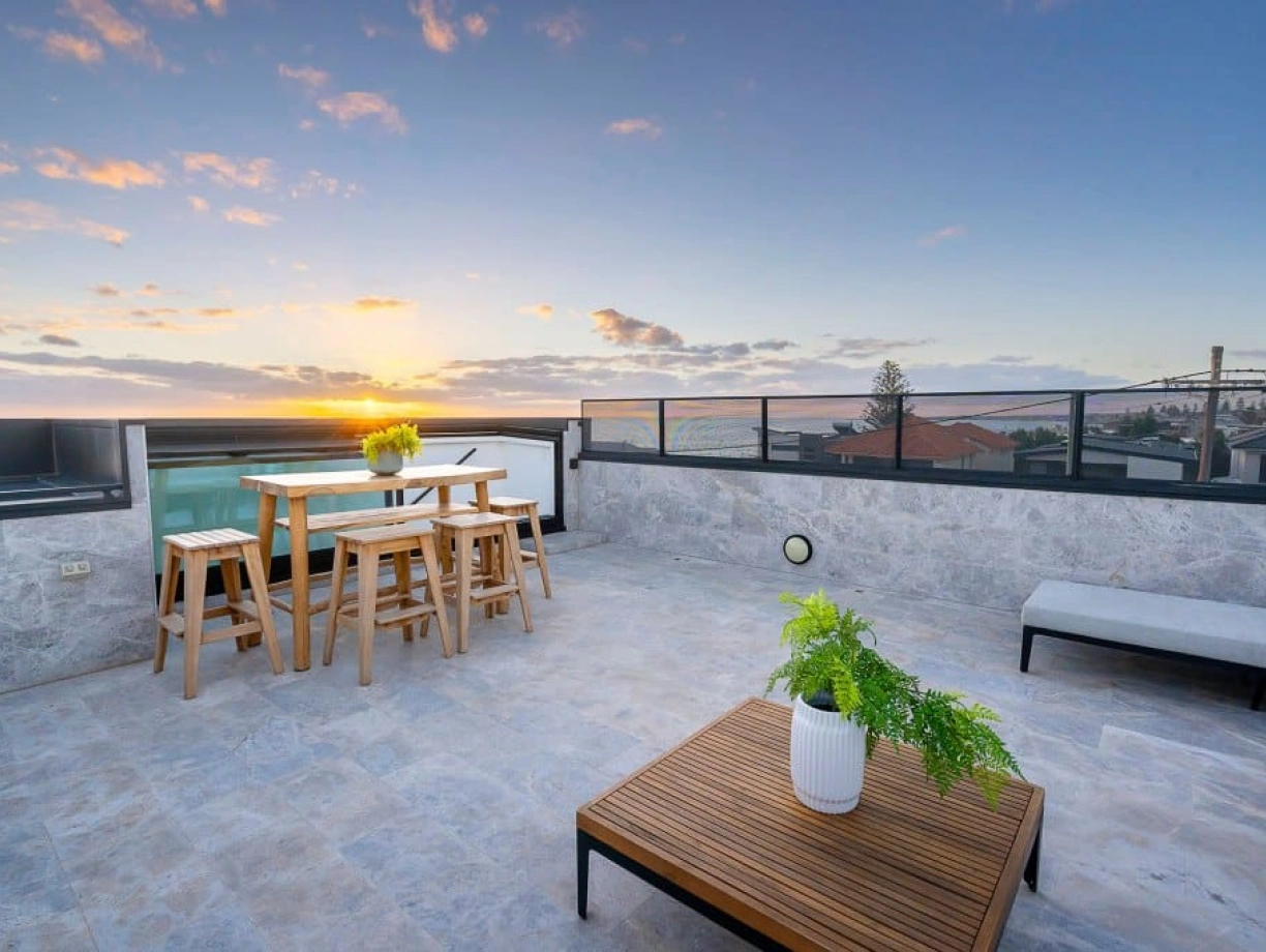 Terraza moderna con una mesa de madera y sillas, iluminada por un atardecer. Hay un sofá y una planta en una maceta blanca. El cielo muestra tonos naranjas y rosados.
