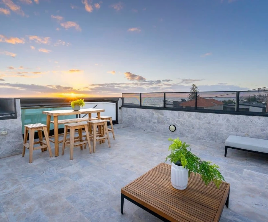 A modern rooftop terrace features a wooden dining set with stools, a potted plant, and a coffee table. The sky is filled with vibrant hues of orange and pink as the sun sets in the background, casting a warm glow over the area.