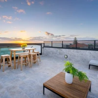 A modern rooftop terrace features a wooden dining set with stools, a potted plant, and a coffee table. The sky is filled with vibrant hues of orange and pink as the sun sets in the background, casting a warm glow over the area.