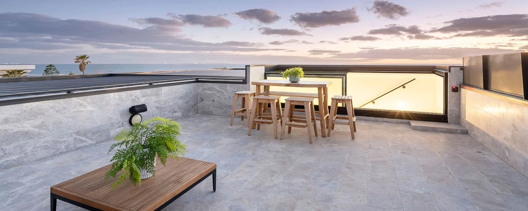Terrasse spacieuse avec mobilier en bois, comprenant une table et des chaises, décorée de plantes. Le coucher de soleil se reflète sur l’horizon, créant une ambiance chaleureuse.