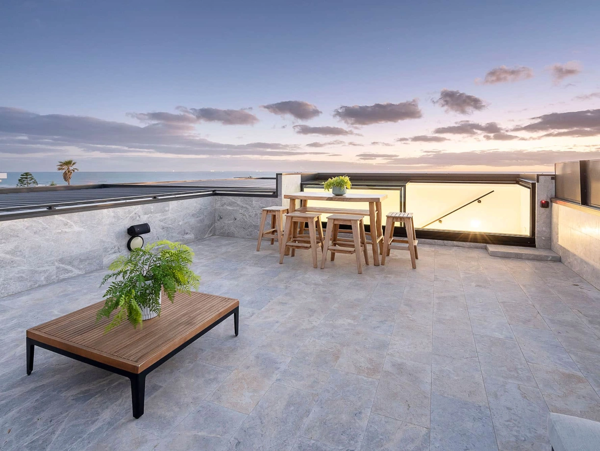 A modern rooftop terrace features a wooden table and stools surrounded by gray stone flooring. Lush green plants add a touch of nature, and the scene is set against a beautiful sunset sky with scattered clouds and a view of the ocean.