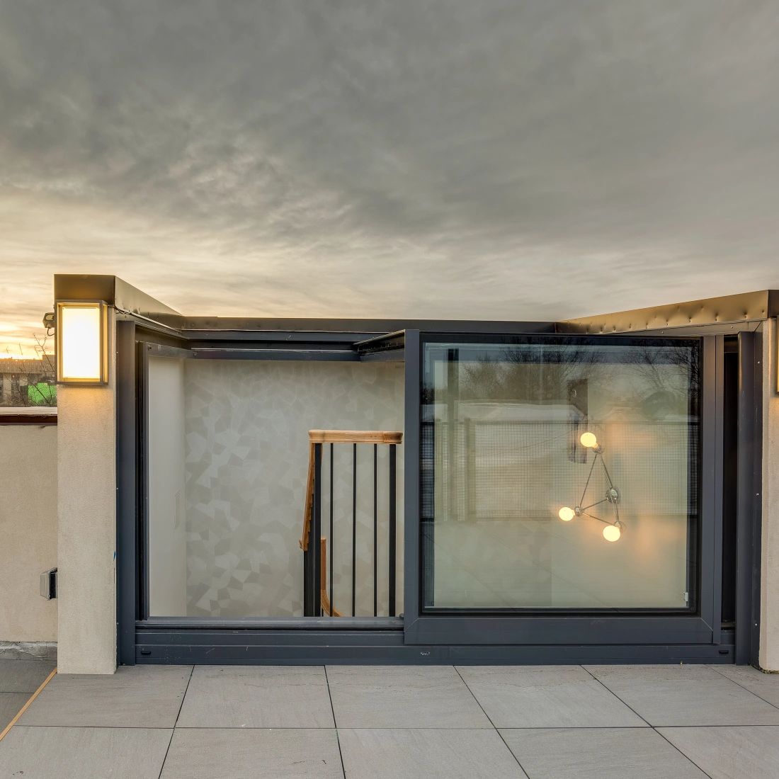 Terraza moderna al atardecer con una entrada a un espacio interior. Faroles de luz en las esquinas y una escalera visible en el interior. Nubes grises en el cielo.