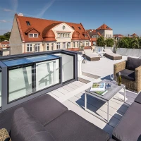 A stylish rooftop terrace features modern outdoor furniture, including a cozy sectional sofa and a small table. The space is surrounded by greenery, with a city skyline and a clear blue sky in the background.