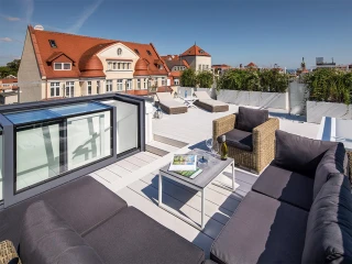 A stylish rooftop terrace features modern gray seating, a small table with a magazine and two glasses, surrounded by greenery and a clear blue sky. In the background, a historic building with a red roof is visible.