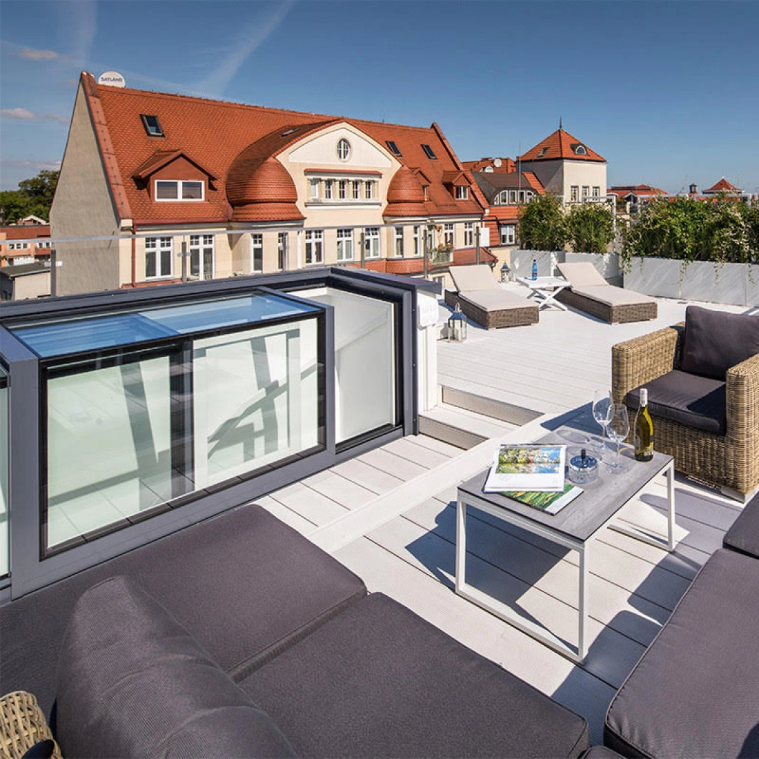 A stylish rooftop terrace features modern outdoor furniture, including a cozy sectional sofa and a small table. The space is surrounded by greenery, with a city skyline and a clear blue sky in the background.