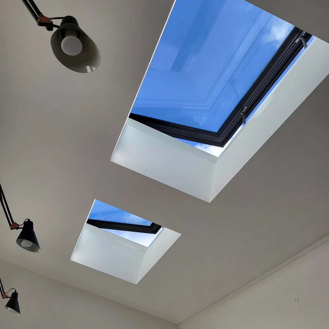Deux fenêtres de toit carrées encadrées de noir laissent entrer la lumière, révélant un ciel bleu parsemé de nuages. Des lampes suspendues au plafond ajoutent de l'éclairage intérieur.