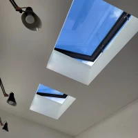 Deux fenêtres de toit carrées encadrées de noir laissent entrer la lumière, révélant un ciel bleu parsemé de nuages. Des lampes suspendues au plafond ajoutent de l'éclairage intérieur.
