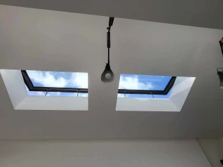 Dos tragaluces en un techo blanco permiten la entrada de luz natural. El cielo azul y algunas nubes son visibles a través de los tragaluces. Hay lámparas colgando en el techo.