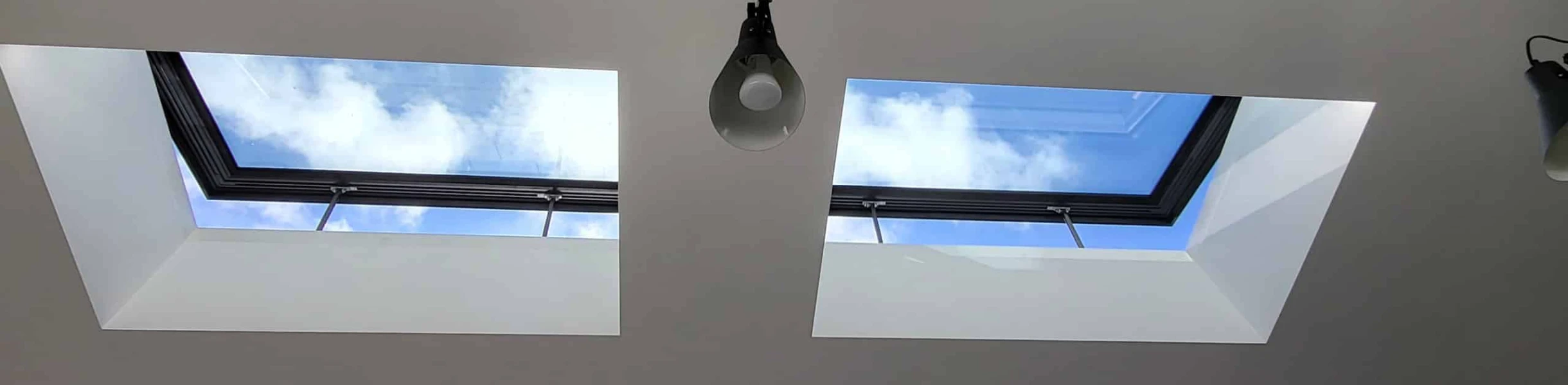 Two skylights in a white ceiling, showing a blue sky with scattered clouds. A single pendant light hangs below one of the skylights.
