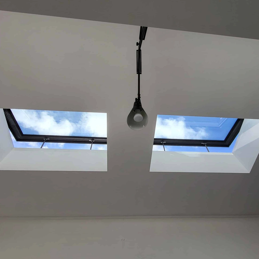Two skylights in a white ceiling, showing a blue sky with scattered clouds. A single pendant light hangs below one of the skylights.