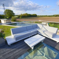 A modern outdoor seating area featuring a gray sectional sofa and a white coffee table on a deck. The background shows a lush landscape with fields, trees, and distant buildings under a partly cloudy sky.