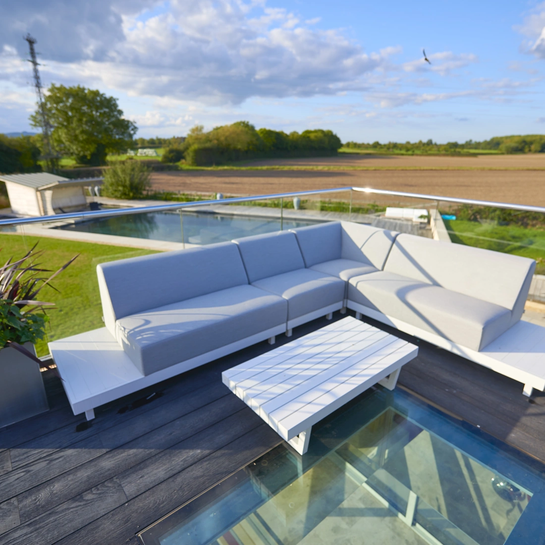 A modern outdoor seating area features a large gray sectional sofa and a white coffee table on a deck with glass panels. Lush greenery and fields extend in the background, under a partly cloudy sky.