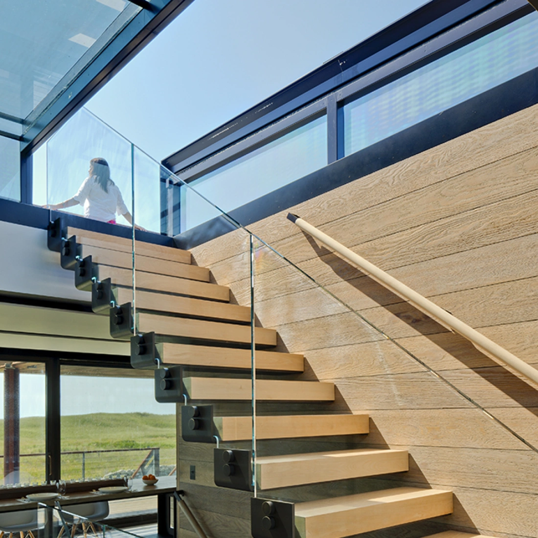 A modern staircase with wooden steps and glass railing, leading to a bright upper floor. Natural light floods the space, highlighting the contemporary design.