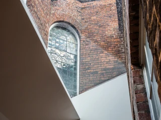 A view of an arched stained glass window set in a brick wall, framed by modern, angular architectural elements. Light filters through the window, creating a contrast between the historic and contemporary design.