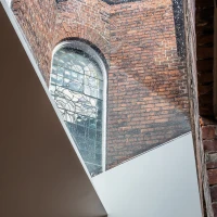 Ein Blick auf ein historisches Fenster aus buntverglastem Glas, eingebettet in rotem Mauerwerk, mit einem modernen architektonischen Element, das diagonal verläuft. Licht strömt durch das Fenster und beleuchtet den Raum.