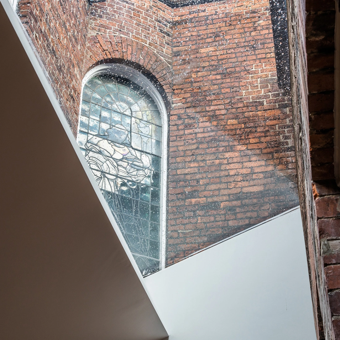 Vista de un espacio arquitectónico donde se combinan ladrillos expuestos y una ventana de cristal colorido. La luz entra a través de la ventana, iluminando las paredes.