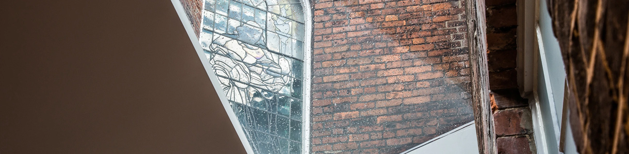 A view of an arched stained glass window set in a brick wall, framed by modern, angular architectural elements. Light filters through the window, creating a contrast between the historic and contemporary design.