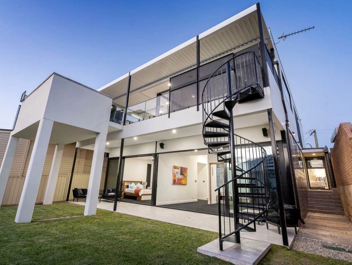 Maison moderne à plusieurs niveaux avec un escalier en colimaçon, un jardin bien entretenu et une verrière donnant sur l’intérieur.